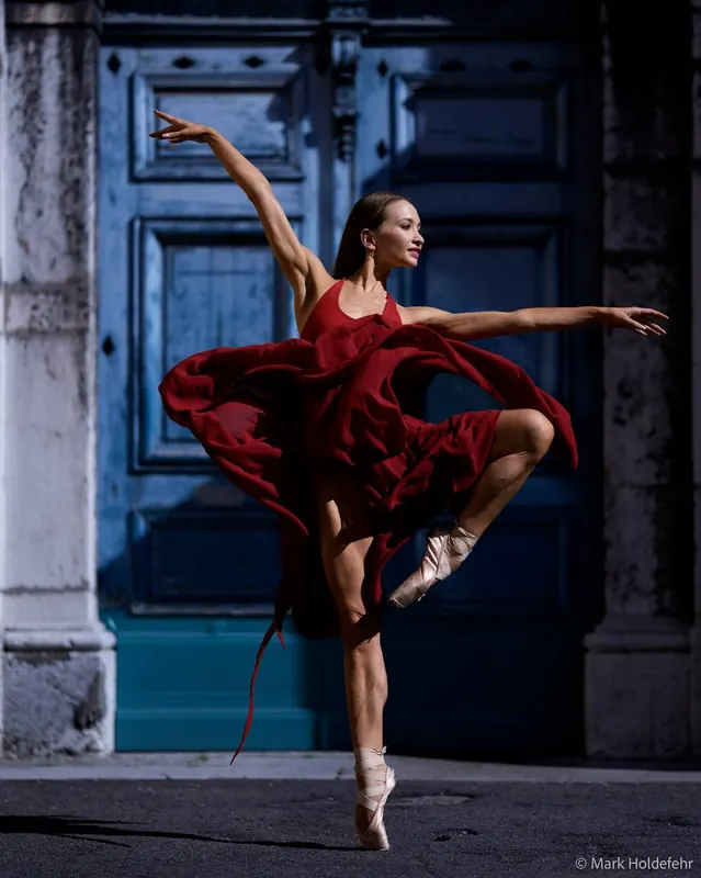 Zuza on pointe in a red dress in front of a blue door, arms open